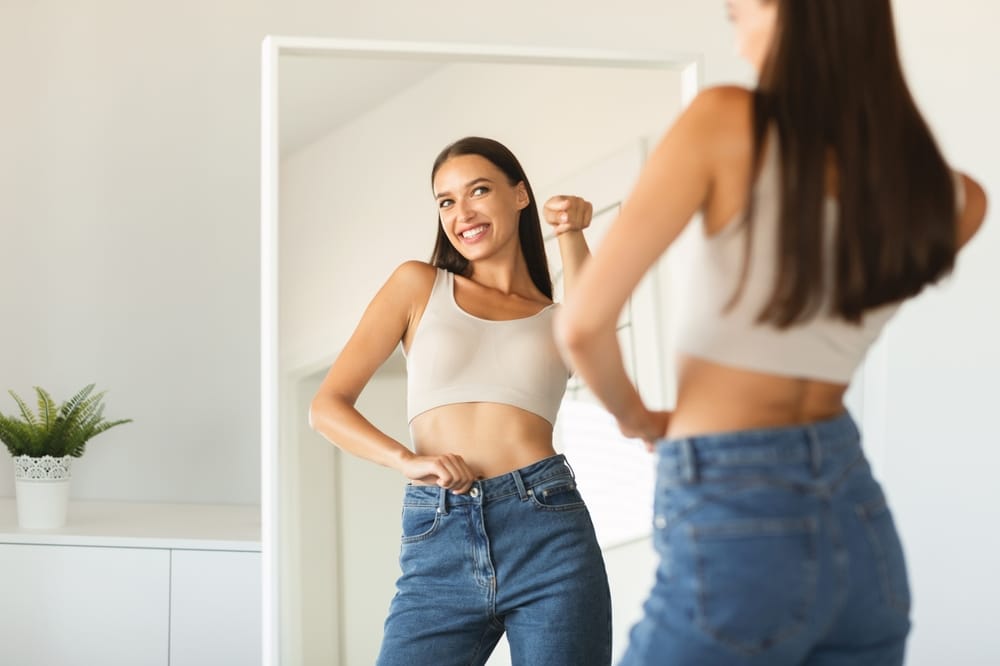 Woman smiling at her reflection while showing progress and improved confidence during a successful weight loss management program – Weight Loss Management